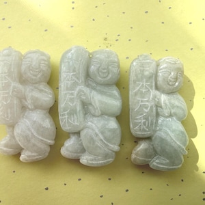 May include: Three jadeite carvings of smiling figures holding tablets with Chinese characters. The carvings are light green and white, with detailed facial features and clothing. The figures are on a yellow background.
