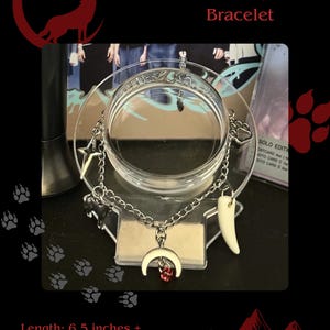 May include: Silver-toned bracelet with a crescent moon charm and a red gem, plus a tooth-shaped charm. The bracelet also features a star and paw print charm. The text reads "&TEAM Inspired Bracelet". The bracelet is 6.5 inches long with a 1-inch extension.