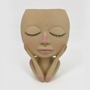 May include: A beige face-shaped planter with closed eyes, black eyelashes, and pink lips and cheeks. The hands are positioned under the chin, with pink fingernails. The planter is a decorative item for plants.