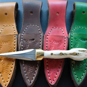 May include: A handcrafted dagger with a bone handle and brass fittings, presented with four leather sheaths in tan, brown, red, and green. The sheaths feature embossed patterns and are stitched with white thread.