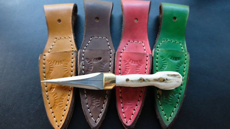 May include: A handcrafted dagger with a bone handle and brass fittings, presented with four leather sheaths in tan, brown, red, and green. The sheaths feature embossed patterns and are stitched with white thread.