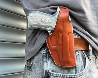 Op de afbeelding: Een zilveren pistool zit in een bruin leren holster, bevestigd aan een riem. Het holster wordt aan de rechterkant gedragen, bevestigd aan een spijkerbroek. Het leren holster heeft een gedetailleerd ontwerp en is gestikt met witte draad.
