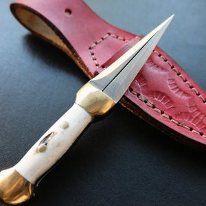 May include: A small, double-edged dagger with a bone handle and brass accents, resting on a red leather sheath. The blade is silver, and the sheath has a decorative embossed pattern.