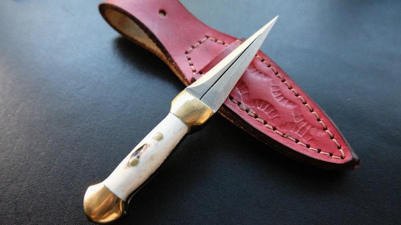 May include: A small, double-edged dagger with a bone handle and brass accents, resting on a red leather sheath. The blade is silver, and the sheath has a decorative embossed pattern.