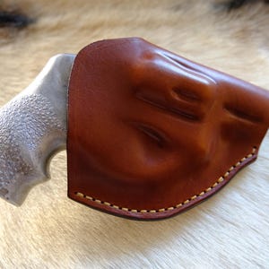May include: A brown leather holster with a stitched border partially covers a silver-colored handgun grip. The holster has a molded design and a rich, glossy finish. The background is a light-colored, textured surface.
