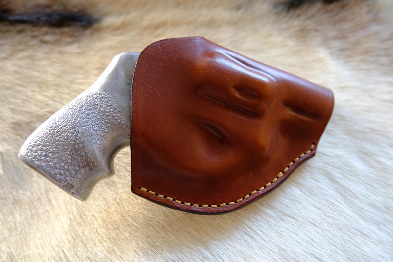 May include: A brown leather holster with a stitched border partially covers a silver-colored handgun grip. The holster has a molded design and a rich, glossy finish. The background is a light-colored, textured surface.