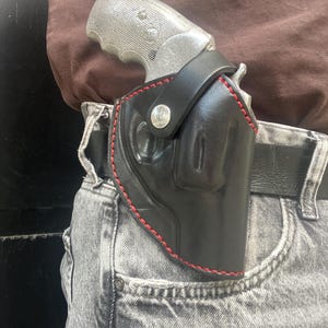 May include: A black leather holster with red stitching and a silver-coloured metal snap button closure. The holster is secured to a black belt and contains a silver-toned revolver with a textured grip, positioned at the waistline of grey jeans.