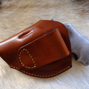 May include: A brown leather holster with a stitched design and a silver-colored gun grip. The holster has a curved shape and a flap. The stitching is done with yellow thread.