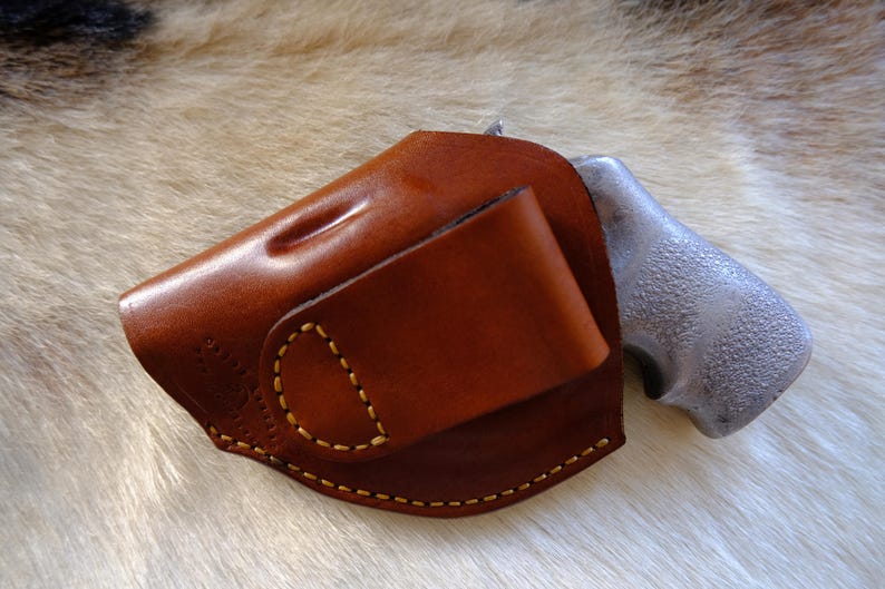 May include: A brown leather holster with a stitched design and a silver-colored gun grip. The holster has a curved shape and a flap. The stitching is done with yellow thread.