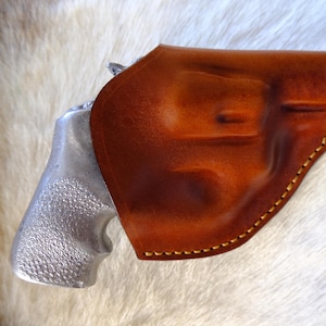 May include: A brown leather holster with a silver-colored handgun. The holster is a rich, warm brown with visible stitching. The handgun's grip is textured. The image is set against a backdrop of white and tan fur.