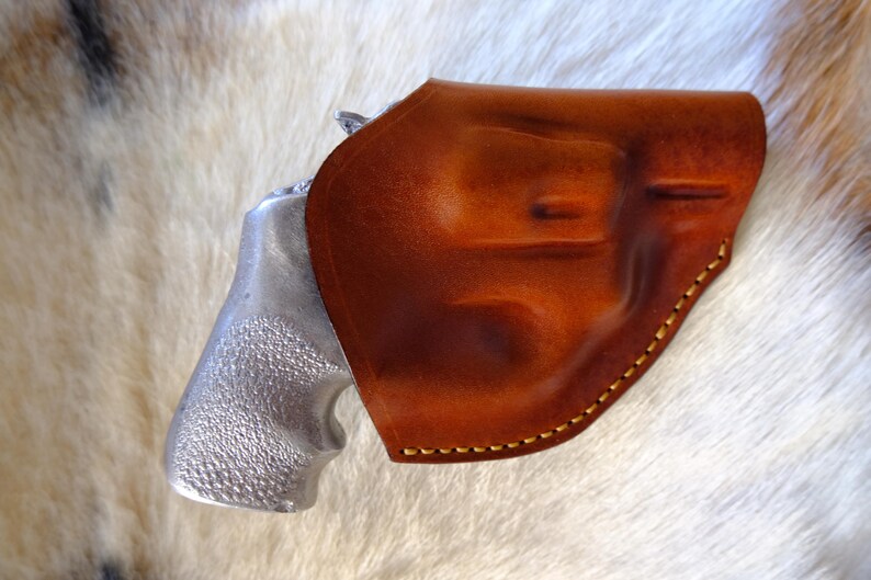 May include: A brown leather holster with a silver-colored handgun. The holster is a rich, warm brown with visible stitching. The handgun's grip is textured. The image is set against a backdrop of white and tan fur.