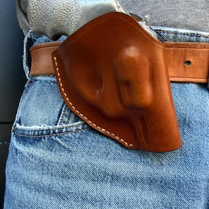 May include: A brown leather holster with white stitching, holding a silver handgun. The holster is attached to a brown leather belt, worn over blue denim jeans. The image is a close-up.