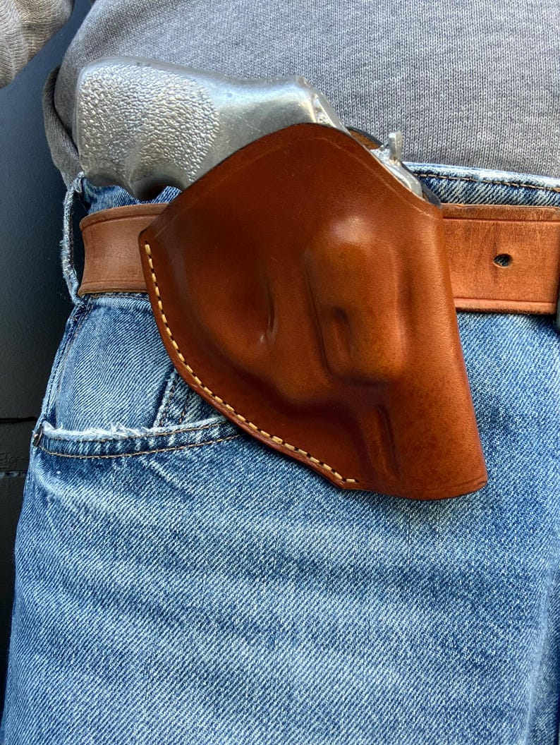 May include: A brown leather holster with white stitching, holding a silver handgun. The holster is attached to a brown leather belt, worn over blue denim jeans. The image is a close-up.