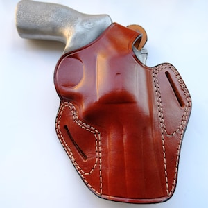 May include: A brown leather holster with white stitching, holding a silver handgun. The holster has a glossy finish and a detailed design, with visible stitching along the edges. The overall theme is related to firearms and accessories.