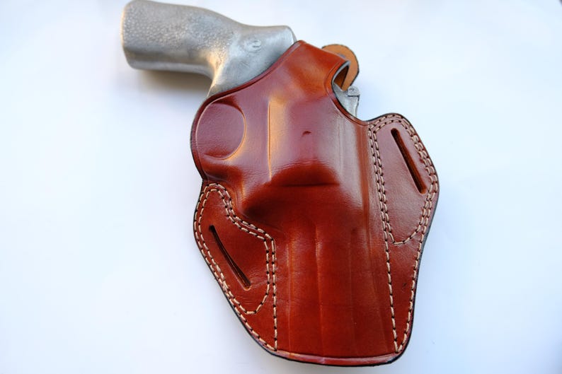 May include: A brown leather holster with white stitching, holding a silver handgun. The holster has a glossy finish and a detailed design, with visible stitching along the edges. The overall theme is related to firearms and accessories.