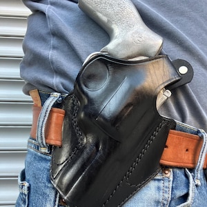 Op de afbeelding: Zwarte leren holster met een zilveren handpistool. De holster wordt gedragen op een bruine leren riem met een blauwe spijkerbroek. De holster is ontworpen om het handpistool veilig vast te houden. De stiksels zijn zichtbaar op de holster.