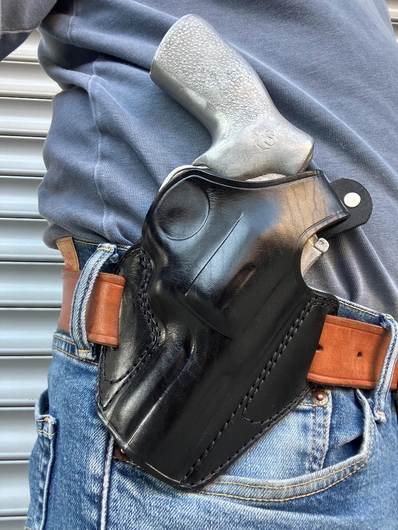 May include: Black leather holster with a silver handgun. The holster is worn on a brown leather belt with blue jeans. The holster is designed to securely hold the handgun. The stitching is visible on the holster.