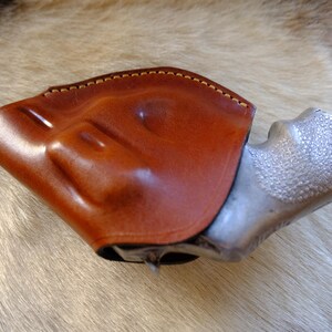 May include: A brown leather holster with a light brown stitched edge, holding a silver-colored handgun. The holster is made of smooth, polished leather. The handgun's grip has a textured surface.