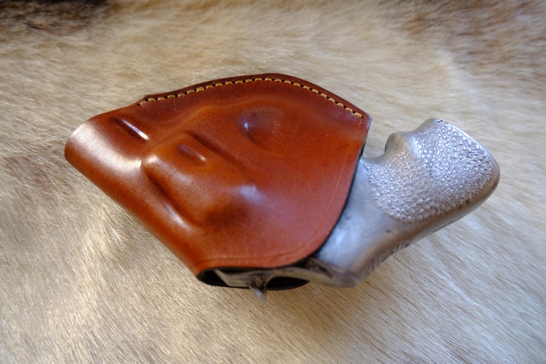 May include: A brown leather holster with a light brown stitched edge, holding a silver-colored handgun. The holster is made of smooth, polished leather. The handgun's grip has a textured surface.