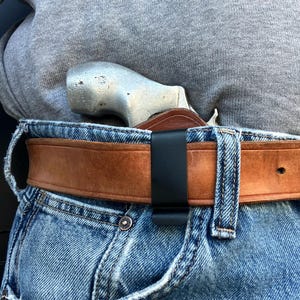 Può includere: Un revolver d'argento è parzialmente visibile, assicurato all'interno di una fondina in pelle marrone, agganciata a una cintura nera. La cintura è indossata con jeans blu. Primo piano della zona della vita.