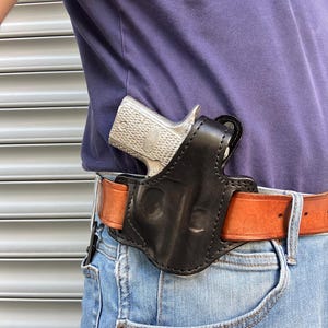 Könnte beinhalten: Ein schwarzes Lederholster mit einer silbernen Pistole darin. Das Holster ist an einem braunen Ledergürtel befestigt.