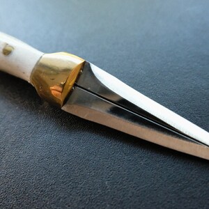 May include: A small, handmade knife with a white handle and brass detailing. The silver blade tapers to a point. The knife is shown against a dark background. The knife's length is approximately 15 cm.