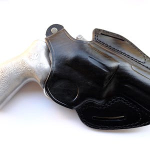 May include: A black leather holster with a silver-colored handgun. The holster is molded to the shape of the gun and features detailed stitching. The handgun's grip is visible, and the overall image is set against a white background.