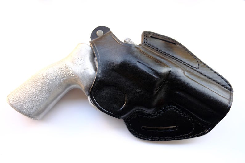 May include: A black leather holster with a silver-colored handgun. The holster is molded to the shape of the gun and features detailed stitching. The handgun's grip is visible, and the overall image is set against a white background.