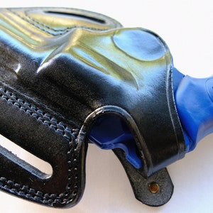 Pode incluir: Um coldre de couro preto com um rev&oacute;lver azul dentro. O coldre tem um passador de cinto e um fecho de press&atilde;o.