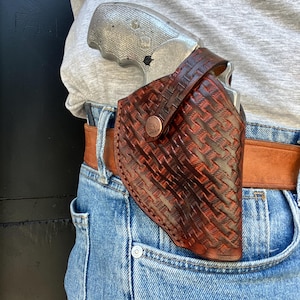 Op de afbeelding: Een donkerbruine lederen holster met een mandvlechtpatroon en een koperkleurige drukknoopsluiting. De holster is bevestigd aan een beige lederen riem en bevat een zilverkleurige revolver met een getextureerde greep, gedragen over een blauwe spijkerbroek.