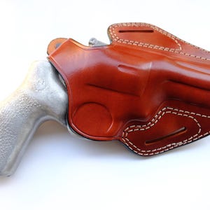 May include: A brown leather holster with white stitching, designed to hold a revolver. The holster is molded to the shape of the gun, with a polished finish. The revolver has a silver-colored handle.