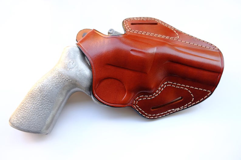 May include: A brown leather holster with white stitching, designed to hold a revolver. The holster is molded to the shape of the gun, with a polished finish. The revolver has a silver-colored handle.