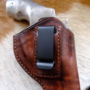 Könnte beinhalten: Eine braune Lederholster mit einem schwarzen Metallclip, zur Aufnahme einer Schusswaffe. Die Holster ist mit einem Kontrastfaden genäht und weist Gebrauchsspuren auf. Die Schusswaffe ist teilweise sichtbar.