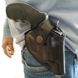 Puede incluir: Una funda de cuero negra con una pistola plateada en su interior. La funda se lleva en la cintura de una persona.
