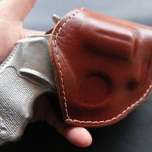 Op de afbeelding: Een zilveren revolver met een bruine leren holster. Het pistool heeft een getextureerde grip en is gedeeltelijk zichtbaar in de holster.