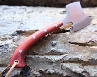 Può includere: Una piccola accetta manuale con un manico in legno curvo color marrone rossiccio, fissato con tre rivetti color argento. La testa dell'ascia in metallo ha una finitura lucida e una lama curva. Un cordino in pelle chiara passa in un occhiello.