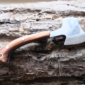 Könnte beinhalten: Ein kleiner, silberner Axt-Schlüsselanhänger mit einem braunen Holzgriff. Die Axt ist an einer braunen Lederschnur befestigt.