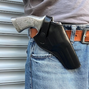 Könnte beinhalten: Eine silberne Handfeuerwaffe in einer schwarzen Lederholster, befestigt an einem braunen Gürtel. Die Holster ist an einer blauen Jeans befestigt.