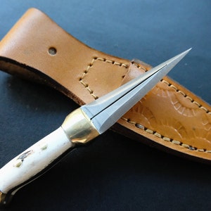 May include: A small, handmade dagger with a polished steel blade and a white handle with brass accents. The dagger rests in a tan leather sheath with decorative stitching.