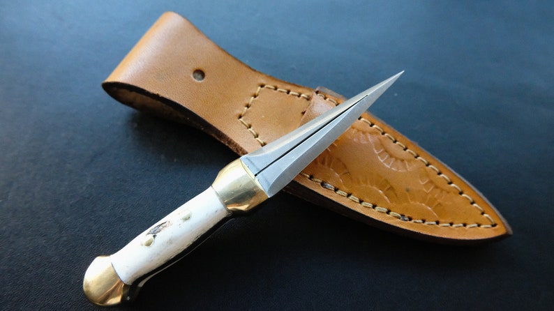 May include: A small, handmade dagger with a polished steel blade and a white handle with brass accents. The dagger rests in a tan leather sheath with decorative stitching.