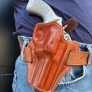 Può includere: Una fondina in pelle marrone con cuciture bianche, che contiene un revolver argentato. La fondina è attaccata a una cintura marrone indossata con jeans blu. L'impugnatura del revolver è visibile.