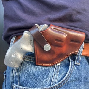 Puede incluir: Una funda de cuero marrón con una correa de cierre a presión, que contiene un revólver de color plateado. La funda está sujeta a un cinturón de cuero marrón sobre unos vaqueros azules. El cuero tiene un acabado liso y pulido con costuras visibles.