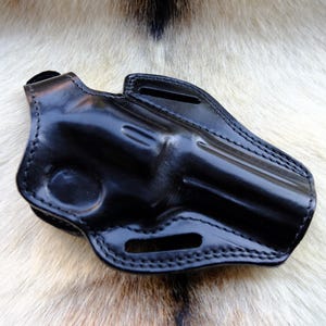 May include: A black leather holster with a glossy finish. The holster is designed to hold a handgun and features stitched detailing along the edges. It has a belt loop and a molded shape for secure firearm storage.