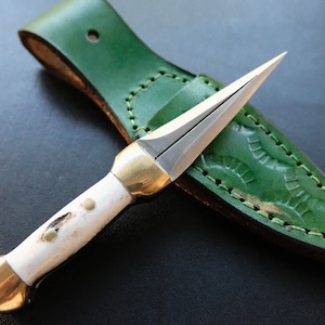May include: A small dagger with a silver blade and a white handle, featuring brass accents. It is presented with a green leather sheath with decorative stitching. The dagger's blade is designed for precision.
