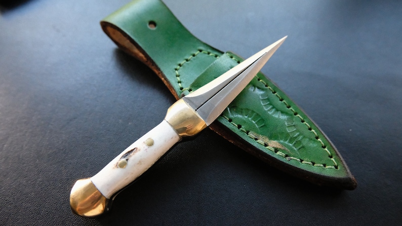 May include: A small dagger with a silver blade and a white handle, featuring brass accents. It is presented with a green leather sheath with decorative stitching. The dagger's blade is designed for precision.