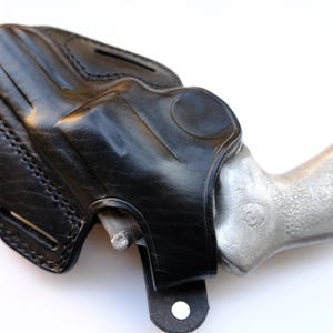 May include: A black leather holster with a silver-colored handgun inside. The holster is stitched and has a loop for a belt. The handgun's grip is textured. The image is set against a white background.