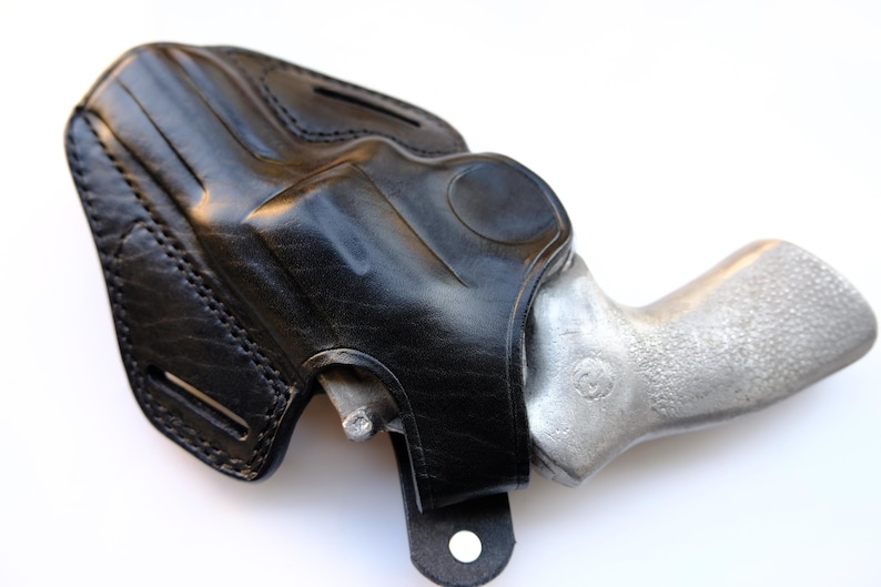 May include: A black leather holster with a silver-colored handgun inside. The holster is stitched and has a loop for a belt. The handgun's grip is textured. The image is set against a white background.