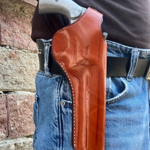 Puede incluir: Una funda de cuero color canela sujeta a un cinturón negro sobre pantalones vaqueros azules, que contiene un revólver de color plateado con empuñadura metálica. La funda presenta contornos moldeados que se ajustan a la forma del arma.