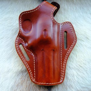 May include: A brown leather holster with white stitching. The holster has a molded design with slots and a loop. The leather has a glossy finish. The item is designed to hold a firearm.