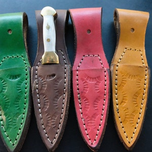 May include: Four small knives with sheaths in green, brown, red, and tan. Each knife has a white handle and a gold-coloured bolster. The sheaths are made of leather with decorative stitching and embossed patterns.